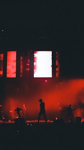 Silhouetted performers on stage with a vibrant red and black background, featuring dynamic lighting and a large screen displaying abstract visuals.
