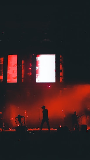 Silhouetted performers on stage with a vibrant red and black background, featuring dynamic lighting and a large screen displaying abstract visuals.