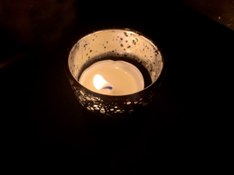 A lit candle placed inside a decorative holder with a perforated pattern, creating a soft glow. Shadows are cast around the candle, enhancing the contrast between light and dark.
