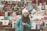 A young person wearing a knit hat and a turquoise jacket stands in front of a colorful market stall. The stall displays a variety of items including knitted hats, scarves, and snacks. Brightly colored packets of chips, soft drinks, and fresh fruit are also visible.