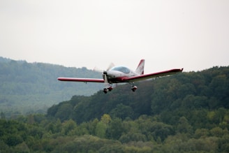 A small aircraft is flying above a lush, green forest-covered landscape. The plane has red accents and a sleek design. The background features rolling hills and a cloudy sky.