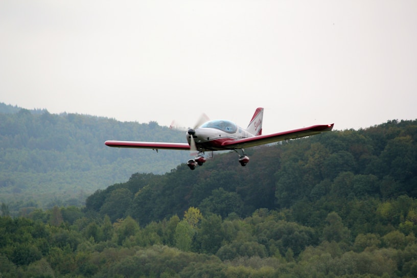 A small aircraft is flying above a lush, green forest-covered landscape. The plane has red accents and a sleek design. The background features rolling hills and a cloudy sky.