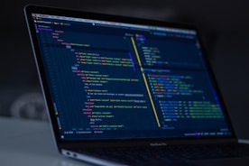 A laptop displays a coding interface with lines of HTML and CSS. The screen shows a dark-themed code editor, possibly for web development, with various tags and attributes visible. The ambient lighting suggests a focused work environment.