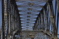 A detailed view of a steel bridge's truss structure showing intricate metalwork and geometric patterns. The perspective looks upward, capturing a clear blue sky through the gaps.