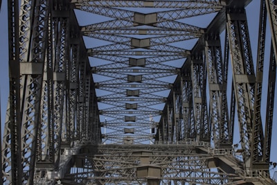 A detailed view of a steel bridge's truss structure showing intricate metalwork and geometric patterns. The perspective looks upward, capturing a clear blue sky through the gaps.