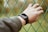 A person's left hand, wearing a black smartwatch, is gripping a green wire fence. The background is blurred with natural green and brown tones, suggesting an outdoor setting.