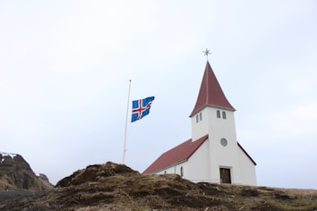 A picturesque white church with a red roof stands prominently on grassy terrain. Next to it, an Icelandic flag waves on a half-raised flagpole. The background features a clear, pale sky and mountains with patches of snow.