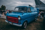 A vintage blue van with red accents is parked on grass, surrounded by people at an outdoor event. The van's classic design includes round headlights and a distinctive grille. Behind the van, there are other vehicles, a group of people, and a large aircraft, suggesting an airfield or car show setting. The sky is overcast, adding a moody atmosphere.