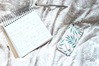 A spiral-bound notebook with handwritten notes lies open on a patterned fabric, accompanied by a pen and a smartphone with a floral case. The fabric's design features intricate paisleys and elegant swirls in muted pastel tones.