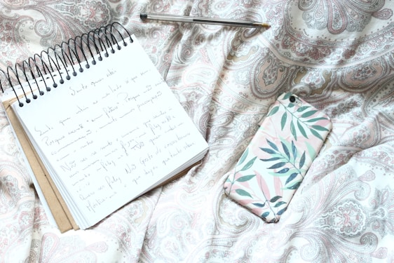 A spiral-bound notebook with handwritten notes lies open on a patterned fabric, accompanied by a pen and a smartphone with a floral case. The fabric's design features intricate paisleys and elegant swirls in muted pastel tones.