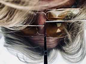 A close-up image of a person's face reflected in a mirror, wearing yellow-tinted sunglasses. The perspective creates an upside-down and somewhat distorted view. The person's hair is light-colored and appears slightly messy.