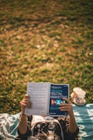 A person is lying on a blanket on grass, holding and reading a book titled 'Understanding Digital Marketing'. Next to the person, a cup from a fast food restaurant can be seen.
