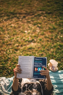 A person is lying on a blanket on grass, holding and reading a book titled 'Understanding Digital Marketing'. Next to the person, a cup from a fast food restaurant can be seen.