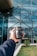 A hand is holding a camera lens in front of a modern glass building. The building features an architectural design with a grid of reflective windows and metal beams. There is greenery in the foreground with small plants and grass.