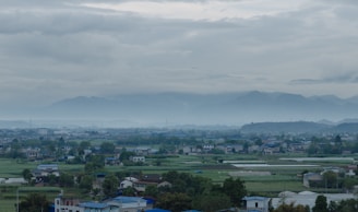A peaceful rural landscape with small houses scattered among vast green fields under a cloudy sky. Distant mountains are faintly visible through the mist, adding to the serene atmosphere.