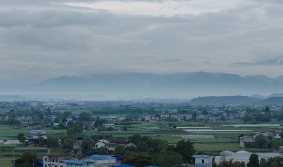 A peaceful rural landscape with small houses scattered among vast green fields under a cloudy sky. Distant mountains are faintly visible through the mist, adding to the serene atmosphere.