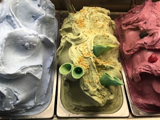 Three tubs of brightly colored gelato are displayed in a showcase. The flavors include blue, green, and pink. The green gelato is garnished with two green cones and sprinkled with chopped nuts. A raspberry and a blackberry are visible on top of the pink gelato.
