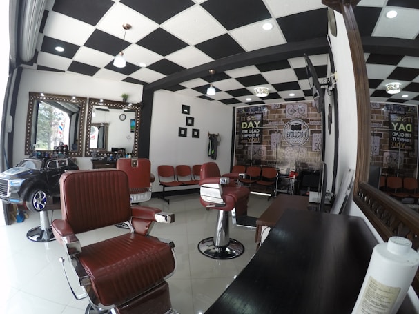 A modern barbershop interior featuring checkered ceiling tiles, several styling chairs with red upholstery, and a row of seating for waiting clients. Large mirrors are mounted on the wall, and a toy car is placed near the window, indicating a family-friendly atmosphere. The walls are decorated with vintage-style posters, adding a retro vibe.