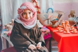 A child wearing a traditional red and white keffiyeh and a brown robe smiles warmly while seated indoors. In the background, various traditional pottery and woven baskets are displayed on red and floral-patterned cloth-covered tables.