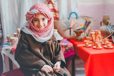 A child wearing a traditional red and white keffiyeh and a brown robe smiles warmly while seated indoors. In the background, various traditional pottery and woven baskets are displayed on red and floral-patterned cloth-covered tables.
