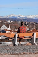 A person wearing a red plaid shirt sits on a bench overlooking a town with rooftops of various shades of orange and brown. In the background, snow-capped mountains stretch across the horizon under a clear blue sky.