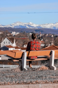 A person wearing a red plaid shirt sits on a bench overlooking a town with rooftops of various shades of orange and brown. In the background, snow-capped mountains stretch across the horizon under a clear blue sky.