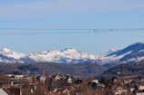 Snow-covered mountains form a majestic backdrop to a small town nestled in the foreground. The town features various rooftops and buildings with tree branches scattered around. Power lines stretch across the view, dividing the sky from the landscape.