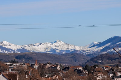 Snow-covered mountains form a majestic backdrop to a small town nestled in the foreground. The town features various rooftops and buildings with tree branches scattered around. Power lines stretch across the view, dividing the sky from the landscape.