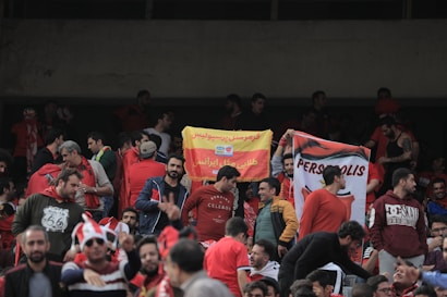 A crowd of people wearing red clothing fills stadium seats. Many are holding banners and flags, with one flag displaying the word 'Perspolis'. The atmosphere appears energetic, likely at a sports event.