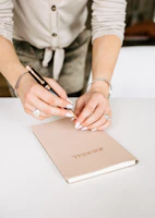 Close-up of a woman journaling with a golden pen on a beige notebook, surrounded by soft fabrics.