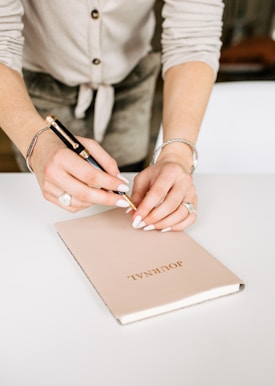 A person writes in a beige journal with the word 'JOURNAL' embossed on the cover using a pen with a gold tip. The individual wears a light-colored cardigan and sports a bracelet and a large ring, highlighting neatly manicured nails. The background is softly blurred, focusing attention on the hands and the journal.