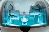 A close-up view of a precision milling machine inside a transparent chamber, likely used for crafting dental prosthetics or intricate mechanical components. The metallic parts are illuminated by a soft blue light, highlighting the machinery’s details and industrial purpose.