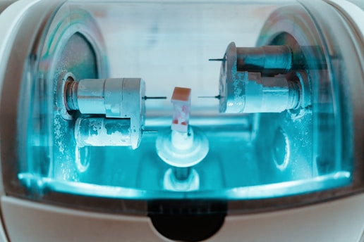 A close-up view of a precision milling machine inside a transparent chamber, likely used for crafting dental prosthetics or intricate mechanical components. The metallic parts are illuminated by a soft blue light, highlighting the machinery’s details and industrial purpose.