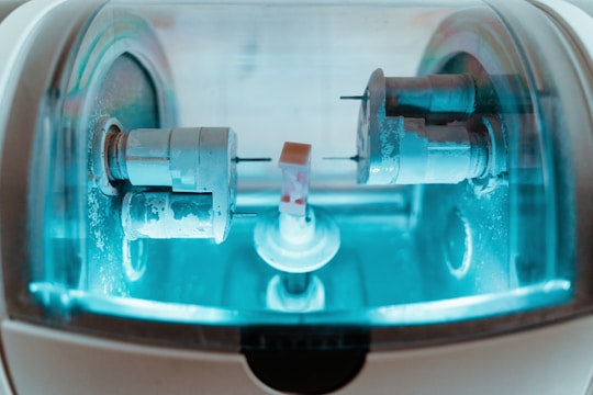 A close-up view of a precision milling machine inside a transparent chamber, likely used for crafting dental prosthetics or intricate mechanical components. The metallic parts are illuminated by a soft blue light, highlighting the machinery’s details and industrial purpose.
