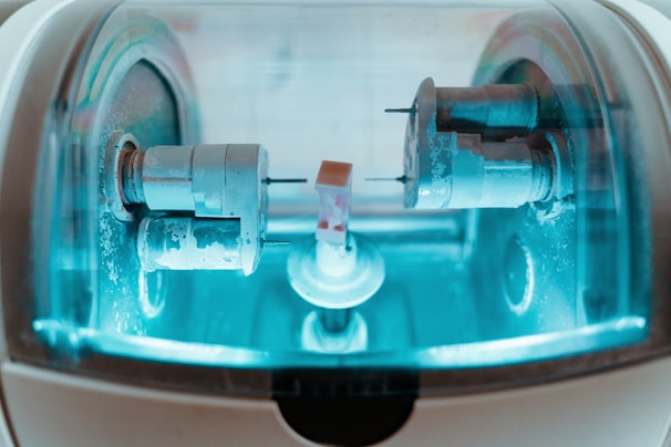 A close-up view of a precision milling machine inside a transparent chamber, likely used for crafting dental prosthetics or intricate mechanical components. The metallic parts are illuminated by a soft blue light, highlighting the machinery’s details and industrial purpose.