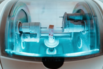 A close-up view of a precision milling machine inside a transparent chamber, likely used for crafting dental prosthetics or intricate mechanical components. The metallic parts are illuminated by a soft blue light, highlighting the machinery&rsquo;s details and industrial purpose.