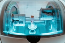 A close-up view of a precision milling machine inside a transparent chamber, likely used for crafting dental prosthetics or intricate mechanical components. The metallic parts are illuminated by a soft blue light, highlighting the machinery&rsquo;s details and industrial purpose.