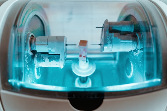 A close-up view of a precision milling machine inside a transparent chamber, likely used for crafting dental prosthetics or intricate mechanical components. The metallic parts are illuminated by a soft blue light, highlighting the machinery&rsquo;s details and industrial purpose.