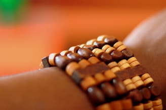 A close-up view of a wrist adorned with a bracelet made of wooden beads arranged in a patterned design. The beads are of varying shades of brown and tan, creating a textured and natural appearance. The background is softly blurred with warm tones of orange and green.