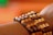 A close-up view of a wrist adorned with a bracelet made of wooden beads arranged in a patterned design. The beads are of varying shades of brown and tan, creating a textured and natural appearance. The background is softly blurred with warm tones of orange and green.