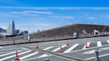 A modern urban landscape featuring a unique architectural structure with slanted, wooden surfaces. The foreground displays road markings and bright orange traffic cones. In the background, a city skyline with distinct skyscrapers can be seen under a bright blue sky.