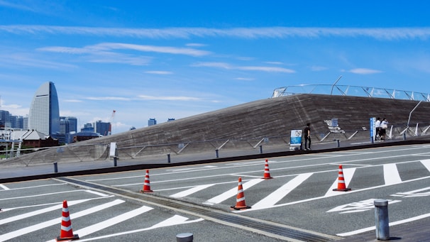 A modern urban landscape featuring a unique architectural structure with slanted, wooden surfaces. The foreground displays road markings and bright orange traffic cones. In the background, a city skyline with distinct skyscrapers can be seen under a bright blue sky.