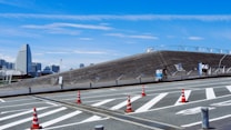 A modern urban landscape featuring a unique architectural structure with slanted, wooden surfaces. The foreground displays road markings and bright orange traffic cones. In the background, a city skyline with distinct skyscrapers can be seen under a bright blue sky.