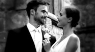 A couple is dressed elegantly, possibly for a wedding. The man is wearing a suit and tie, while the woman is in a sleeveless dress, holding a bouquet. They are gazing at each other affectionately, suggesting a warm and loving connection. The background features a stone wall, indicating an outdoor or historic setting.
