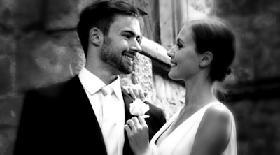 A couple is dressed elegantly, possibly for a wedding. The man is wearing a suit and tie, while the woman is in a sleeveless dress, holding a bouquet. They are gazing at each other affectionately, suggesting a warm and loving connection. The background features a stone wall, indicating an outdoor or historic setting.