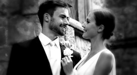 A couple is dressed elegantly, possibly for a wedding. The man is wearing a suit and tie, while the woman is in a sleeveless dress, holding a bouquet. They are gazing at each other affectionately, suggesting a warm and loving connection. The background features a stone wall, indicating an outdoor or historic setting.
