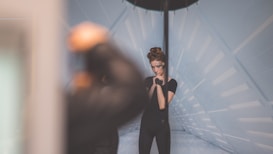 A woman with styled hair and intricate eye makeup is posing in a futuristic setting with geometric patterns on the walls. The focus is on her as she stands beside a large pole, hands together and looking contemplative. The background features soft blue lighting, adding to the modern aesthetic.