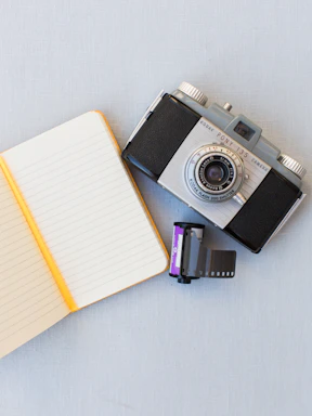 A vintage film camera resting on a wooden table beside a notebook and pen.