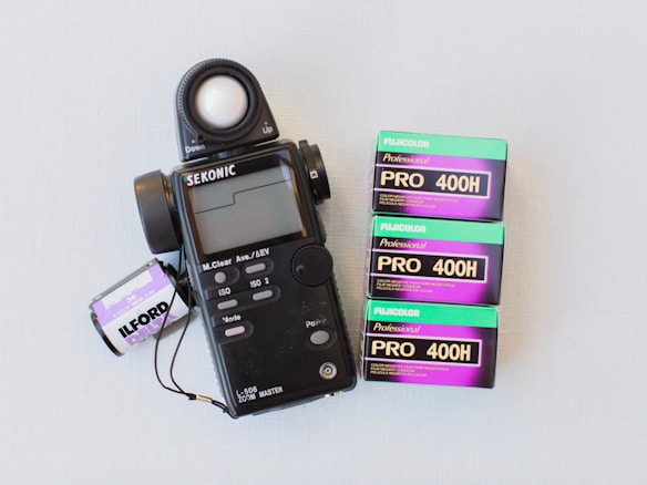 A Sekonic light meter is positioned next to three boxes of Fujicolor Pro 400H film and an Ilford film canister. The light meter has a large spherical sensor and a digital display with various buttons and settings. The Fujicolor boxes are green and purple, indicating they are color negative films with a 400 ISO speed. The Ilford canister is black and white, suggesting a different type of film.