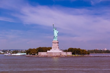 The image features the Statue of Liberty prominently set against a backdrop of a clear blue sky. The iconic statue stands on Liberty Island surrounded by water, with trees and a few buildings visible in the background. A ferry can be seen on the water near the island.